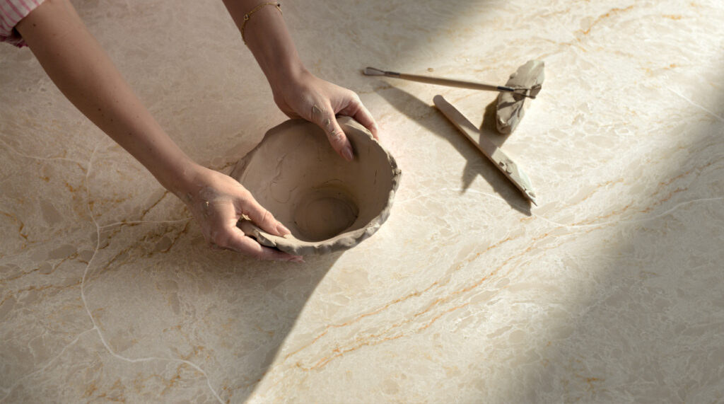 Taj_Whisper_1282x715_RGB-1024x571 Hands shaping clay pottery on a surface, with carving tools nearby in sunlight.